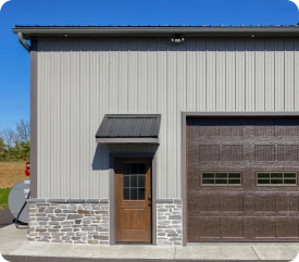 Exterior view of a modern metal building showing a custom wooden entry door, a dark brown garage bay door, and stone veneer wainscoting, built by ISM Construction for Custom Garages in Lebanon, PA.