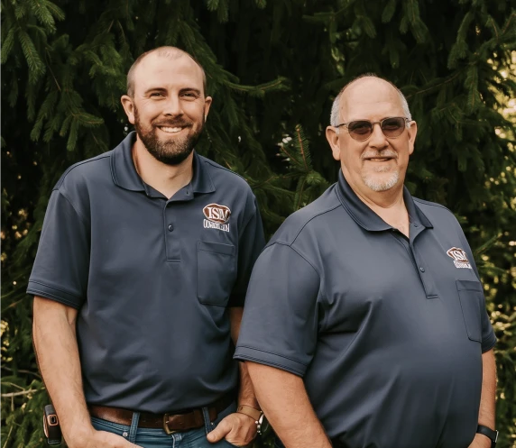 Two ISM Construction team members in navy branded shirts standing outdoors before green pine branches.
